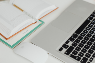 Notebook aberto com teclado visível ao lado de agenda aberta e mouse branco sobre mesa organizada.