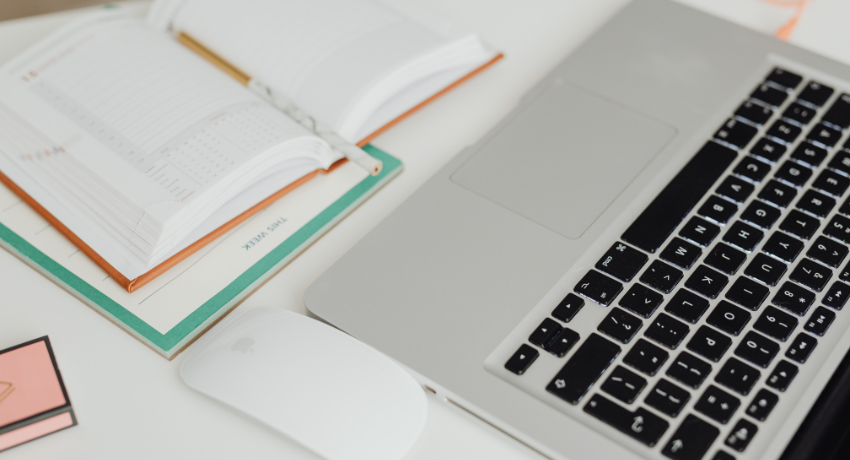 Notebook aberto com teclado visível ao lado de agenda aberta e mouse branco sobre mesa organizada.