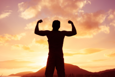 Pessoa em silhueta flexiona os braços em gesto de força contra o pôr do sol alaranjado, com montanhas ao fundo.