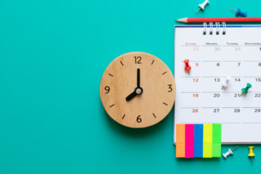 Relógio de parede ao lado de calendário de mesa com datas marcadas e notas adesivas coloridas.