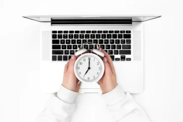 Imagem que ilustra a diferença entre produtividade e eficiência, com uma pessoa segurando um relógio em frente ao teclado de um laptop, destacando o gerenciamento de tempo na prática.